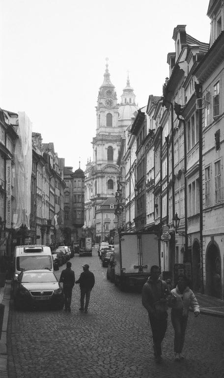 A street in Prague is lined with buildings and cars, with a large church visible in the distance.