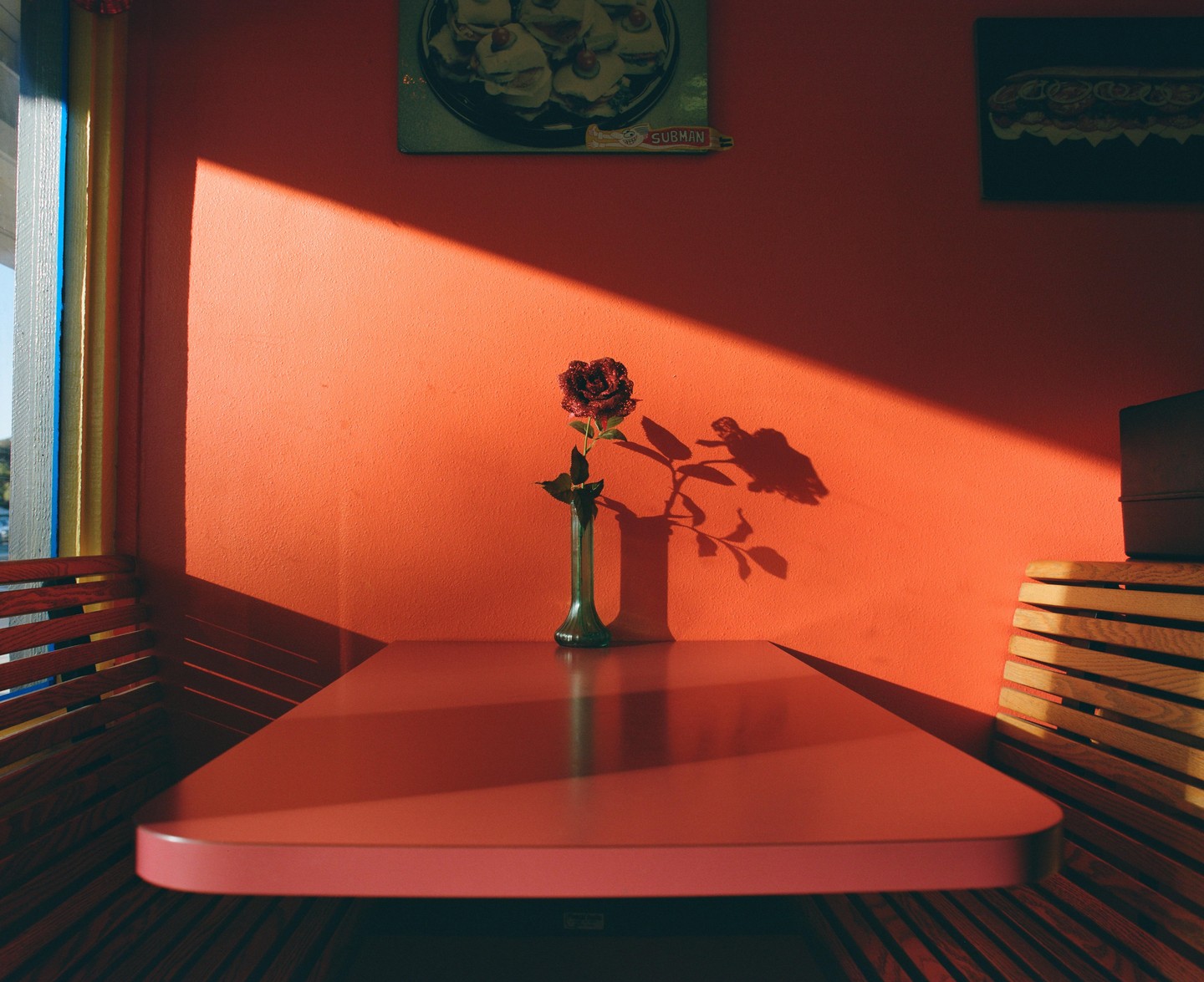 A single rose in a vase sits on a table in a diner booth, with sunlight casting a shadow on the orange wall.