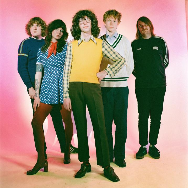 Five musicians pose for a group portrait against a pink and yellow backdrop.