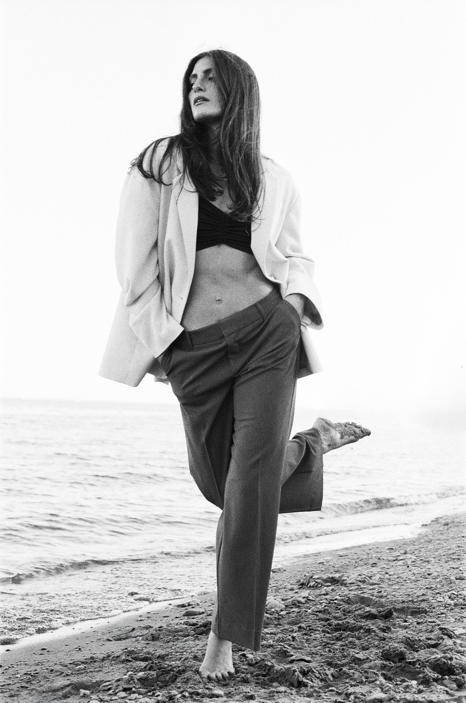A woman stands barefoot on a sandy beach with one foot lifted, wearing a blazer, bra, and trousers.