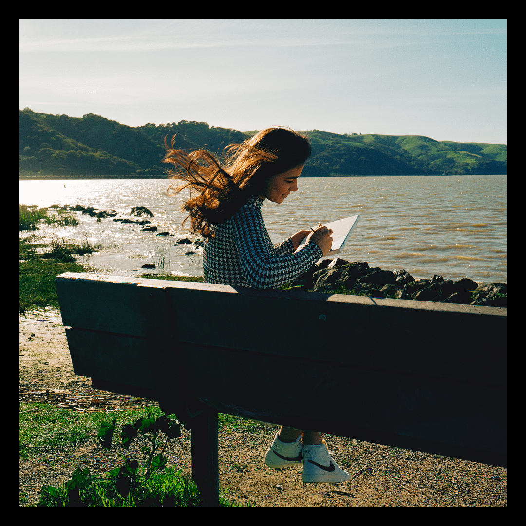 A woman sits on a bench by a body of water, drawing in a sketchbook.