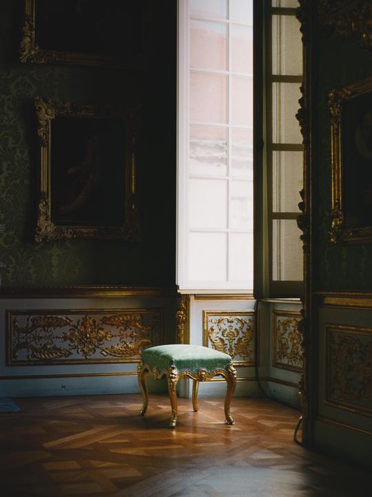 A green velvet stool with golden legs sits on a wooden floor in a room with ornate golden wall decorations and a large window.