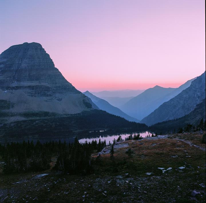 A lake reflects the pink and blue sky at dusk, surrounded by mountains and forest.