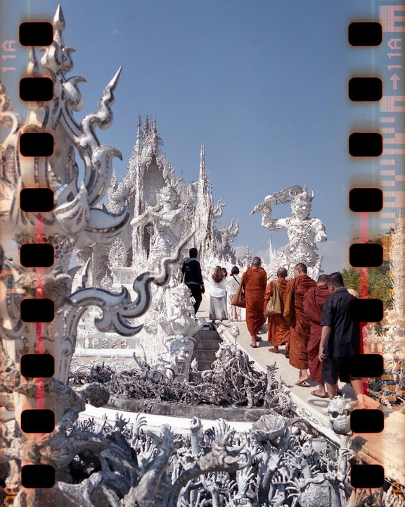 A group of people, including several monks, walk on a path surrounded by white, ornate sculptures under a clear sky.
