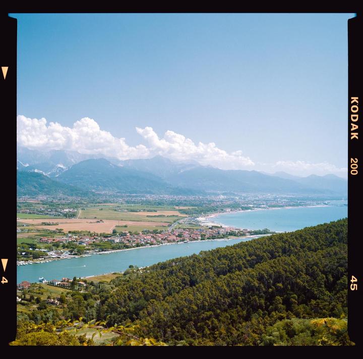 A coastal town with a river and a beach is nestled between a forested hill and distant mountains under a clear sky with scattered clouds.