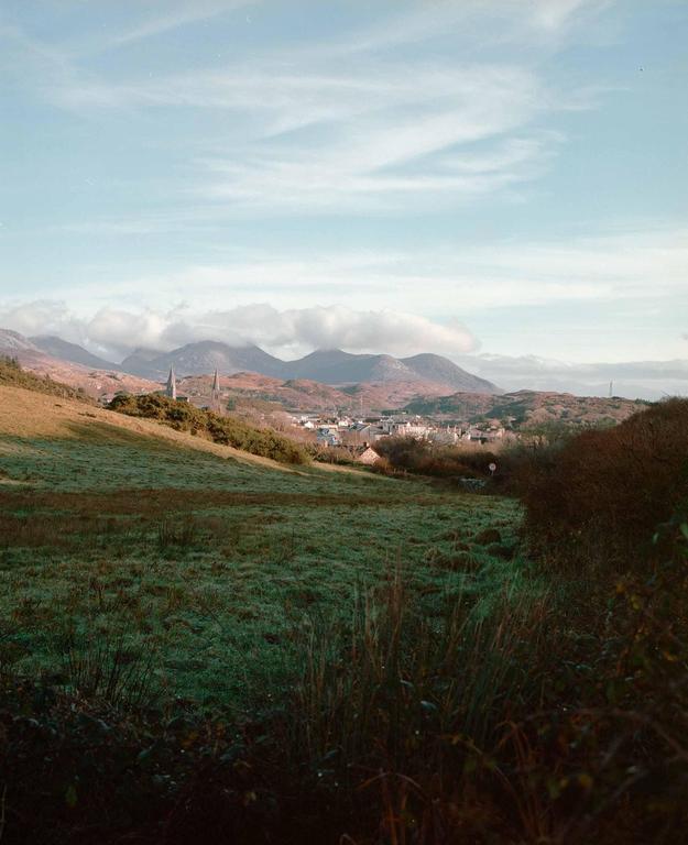 A village with two prominent spires is nestled in a valley, surrounded by rolling hills and mountains under a partly cloudy sky.