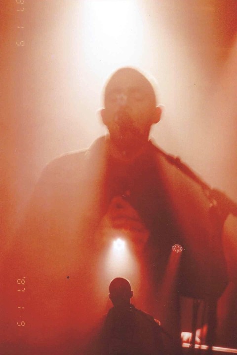 A double exposure shows a man with a bald head in the foreground and a larger, blurry image of a man with a bald head in the background, both illuminated by spotlights.