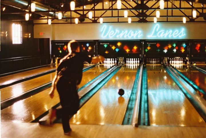 A man bowls a black bowling ball down a lane toward pins under a neon sign that reads "Vernon Lanes."