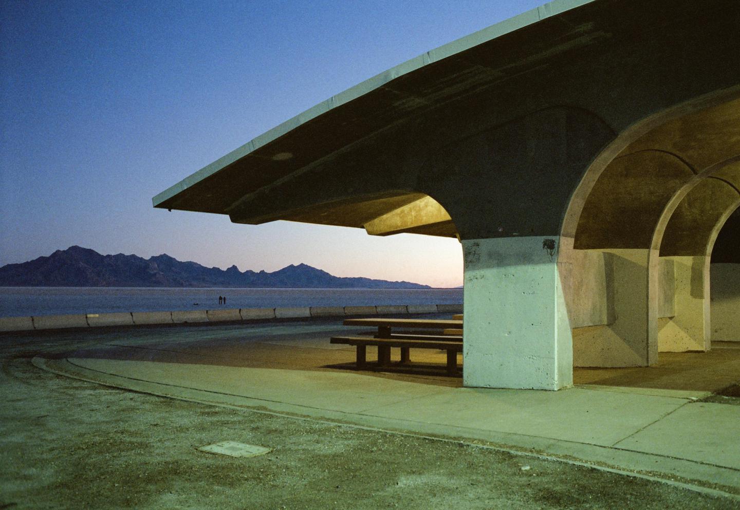 A concrete structure with picnic tables sits beside a body of water with mountains in the distance under a twilight sky.