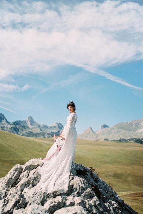 A woman in a white lace dress stands on a rocky outcrop with mountains and a grassy field in the background.
