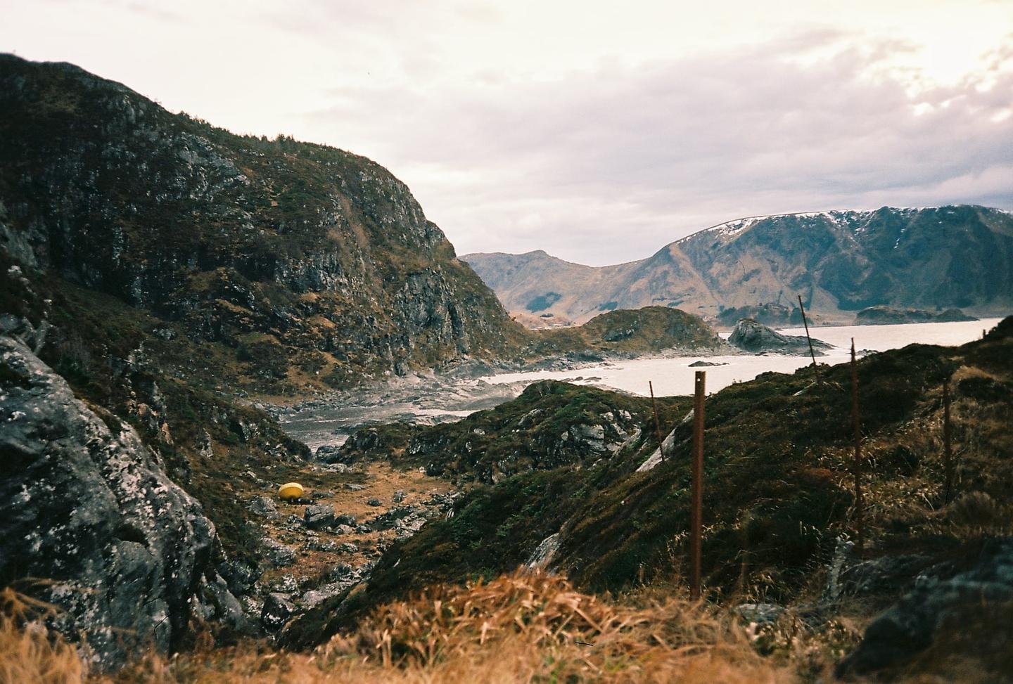 A yellow tent is pitched in a rocky valley leading to a body of water surrounded by mountains.