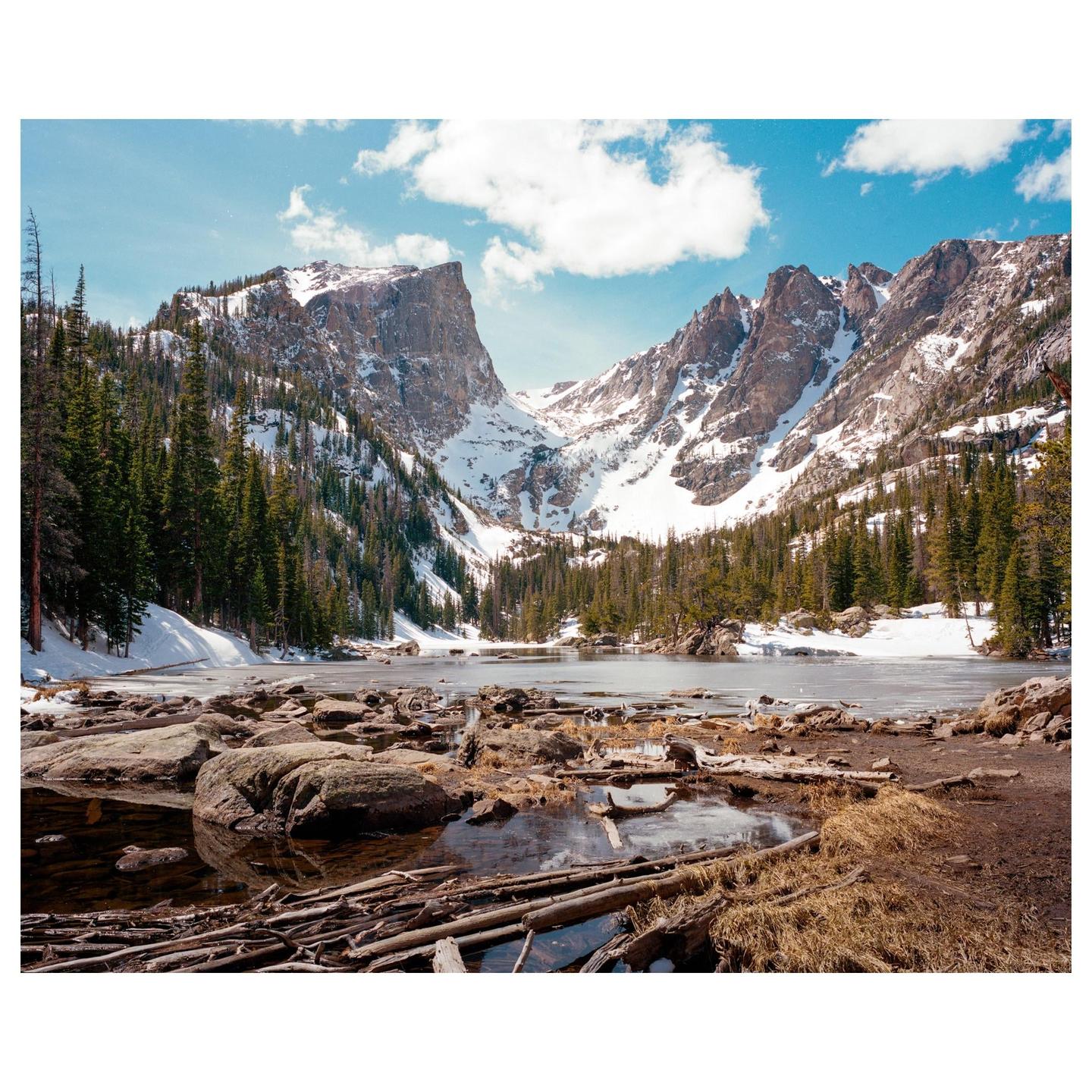 A partially frozen lake is surrounded by snow-covered mountains and pine trees under a blue sky with white clouds.