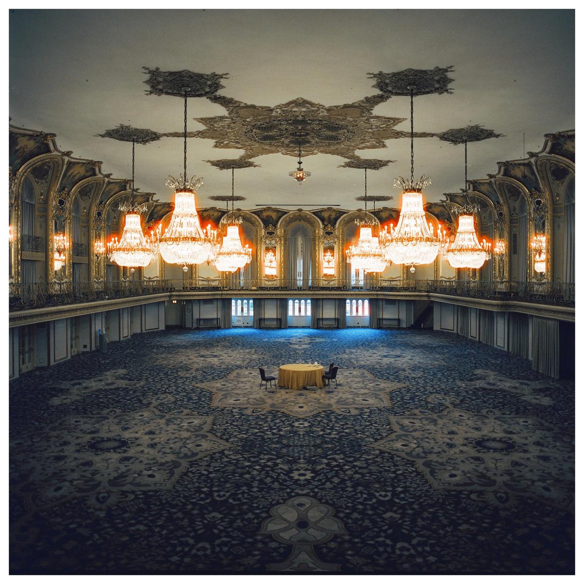 A large, ornate ballroom with many chandeliers has a small table and two chairs in the center of a patterned carpet.