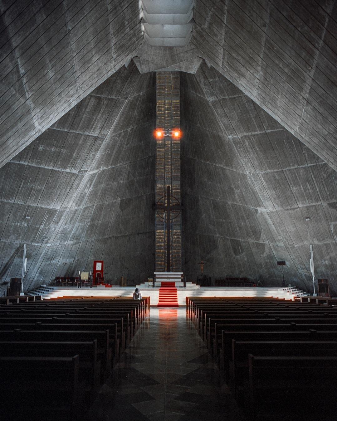A woman sits in a brutalist cathedral with a red carpet leading to an altar.
