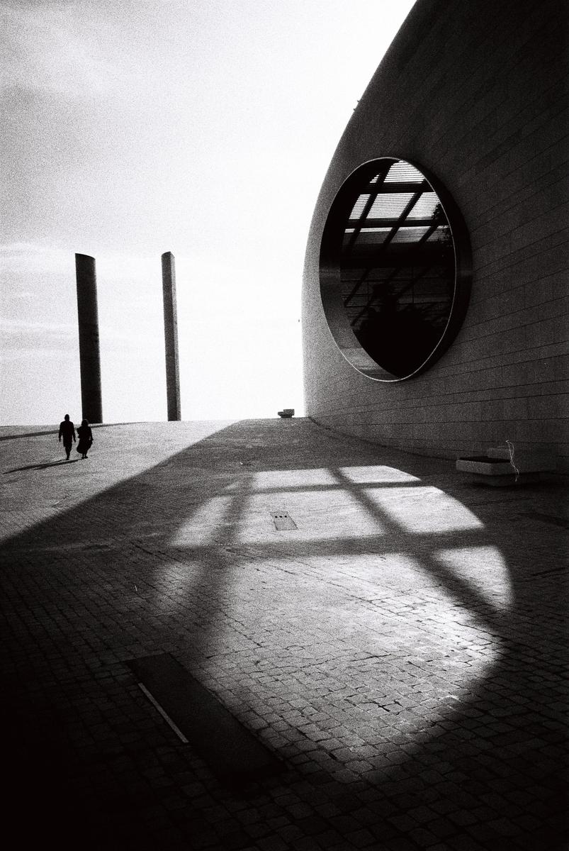Two people walk on a paved surface next to a modern building with a large circular window, casting a grid shadow.