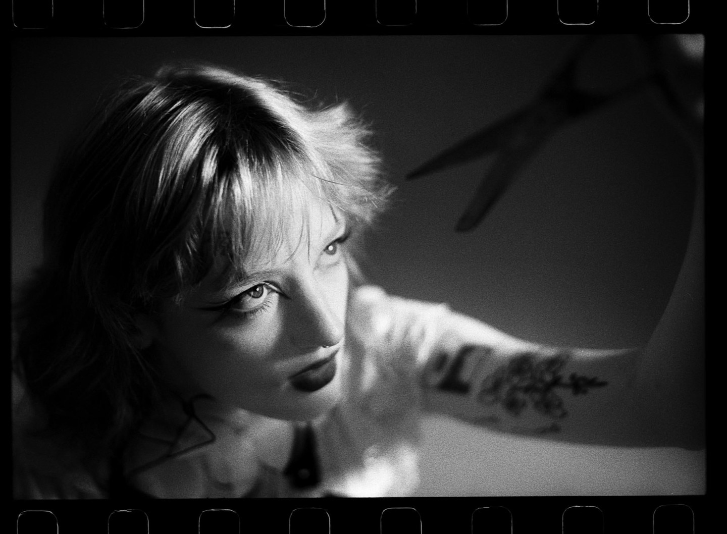 A woman with short hair and a nose ring looks up at a pair of scissors.