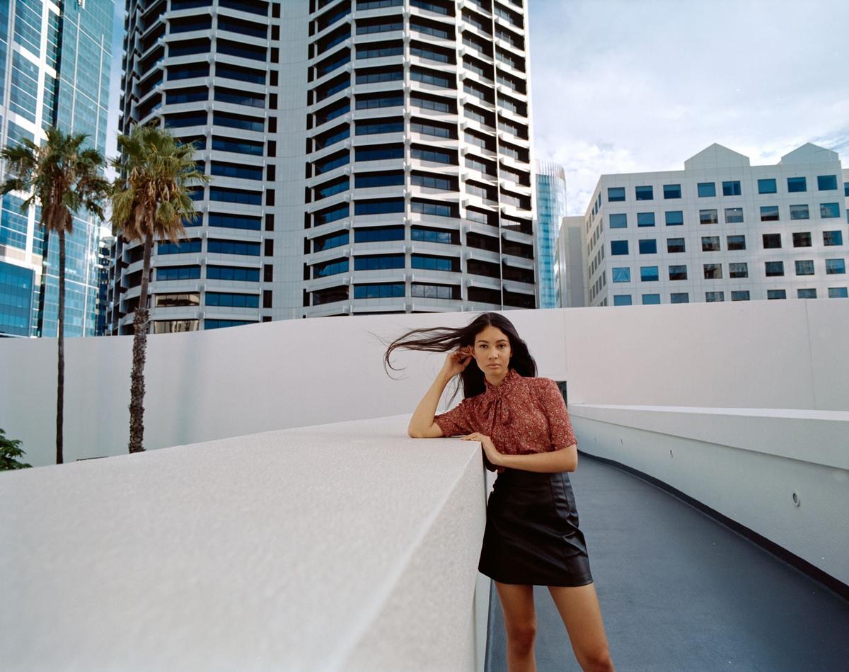 A woman with long dark hair stands on a curved walkway with tall buildings and palm trees in the background.