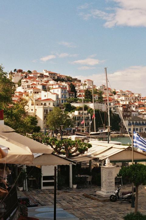 A hillside town with white buildings and red roofs overlooks a harbor with boats and a Greek flag.