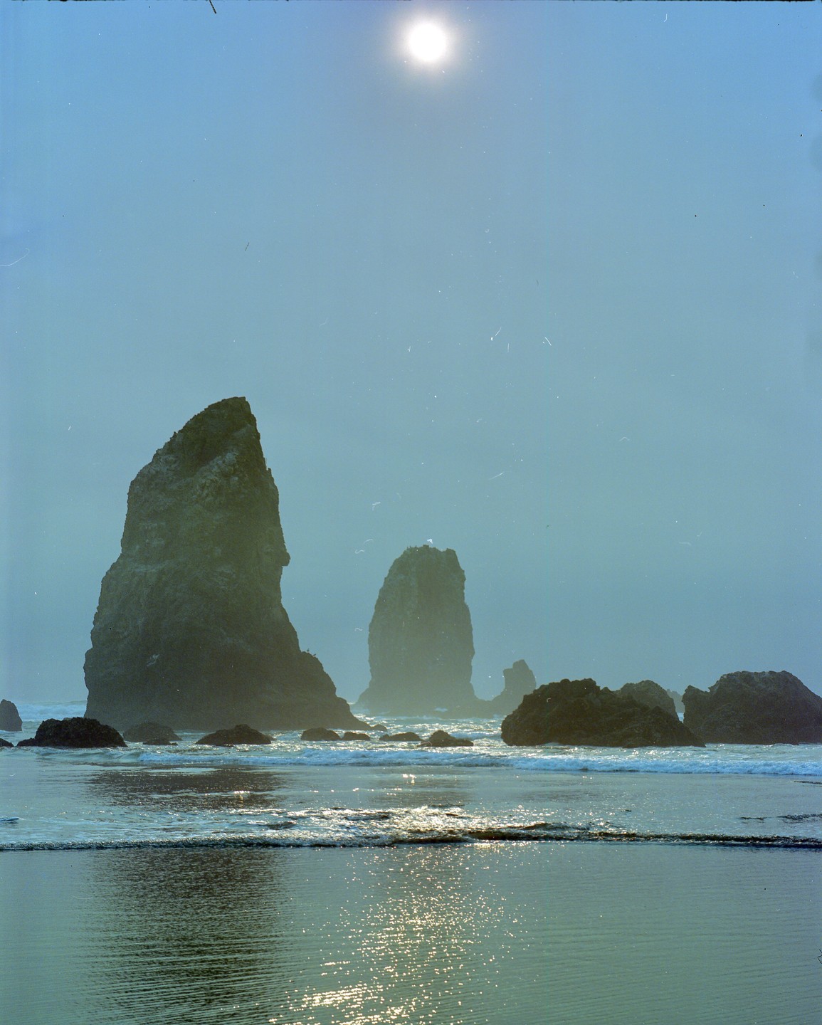 The sun shines brightly over a hazy beach with large sea stacks and gentle waves.