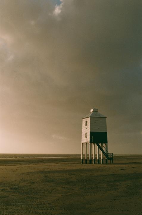 A white and dark green lighthouse stands on stilts on a sandy beach under a cloudy sky.