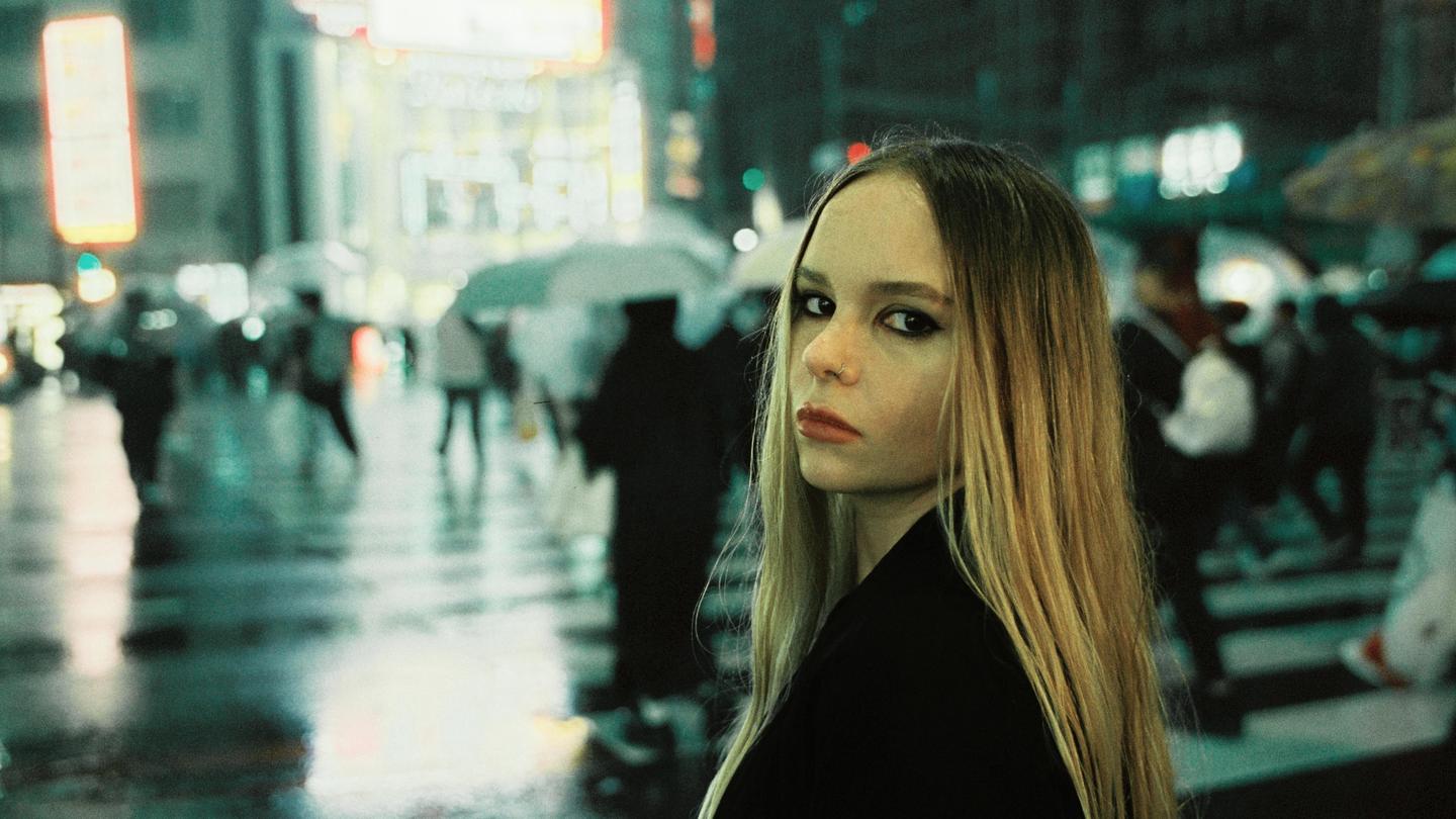 A woman with long hair looks over her shoulder at the viewer on a rainy city street at night.