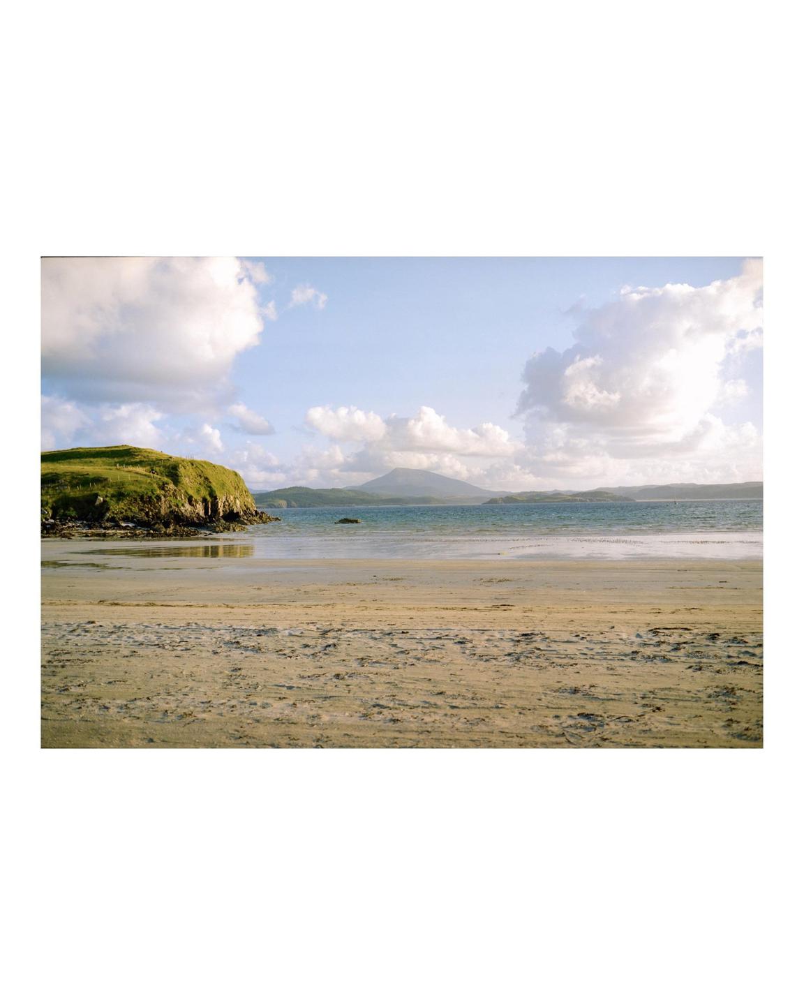 A sandy beach with a grassy hill on the left and a mountain in the distance under a cloudy sky.