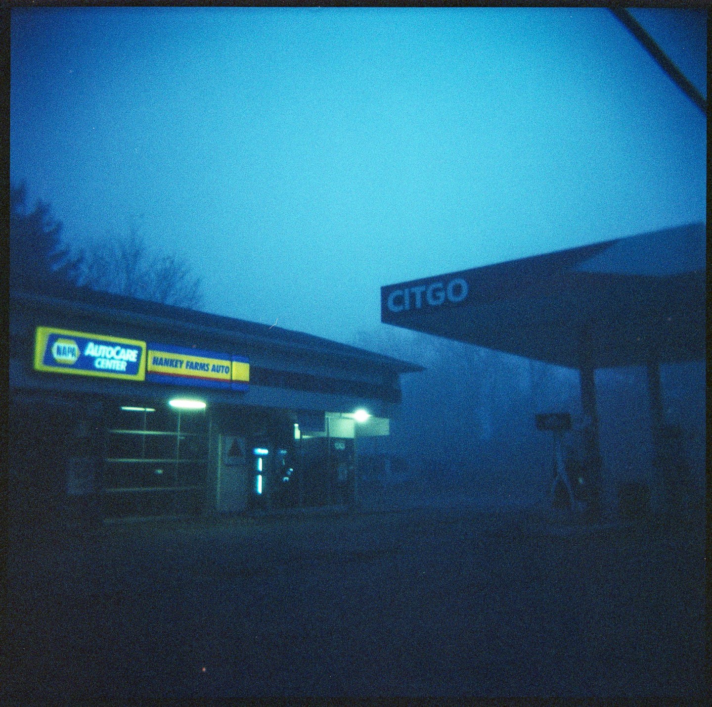 A gas station and auto repair shop are shrouded in blue fog at night.