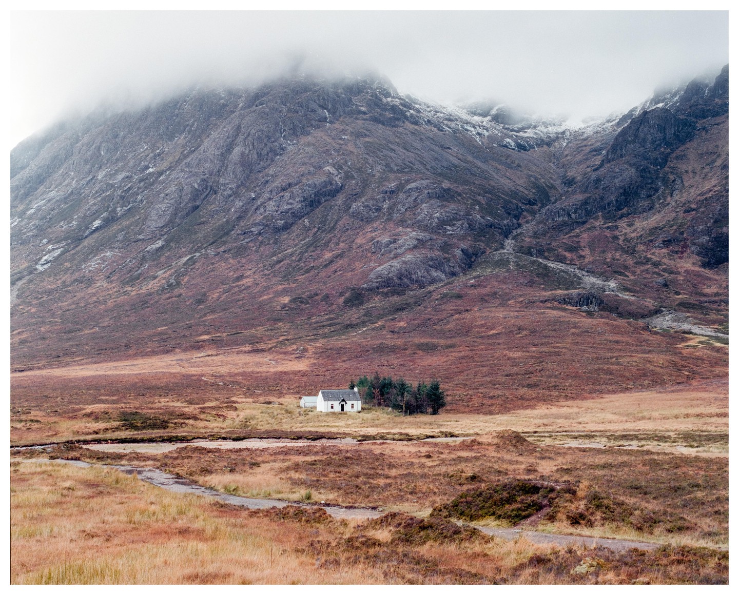 A small white house sits in a vast, rugged landscape with mountains and a winding path.
