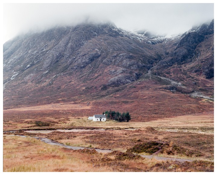 A small white house sits in a vast, rugged landscape with mountains and a winding path.
