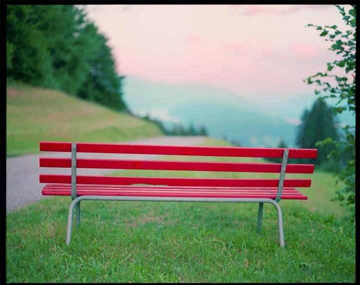A red bench sits on a grassy hill overlooking a mountain landscape with a winding road.