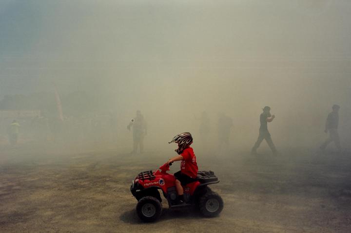 A child in a helmet and red shirt sits on a red toy ATV in a hazy, dusty area with several blurred figures in the background.