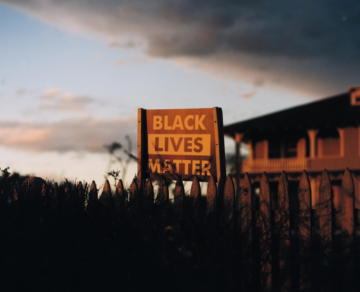 A "Black Lives Matter" sign stands behind a wooden fence with a house and sky in the background.