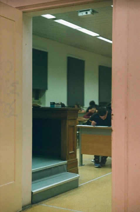 A student sits at a desk taking an exam in a classroom, viewed through a doorway.