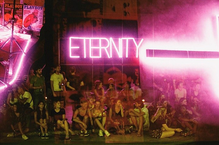 A group of young people are gathered under a bright pink neon sign that reads "ETERNITY" at night.