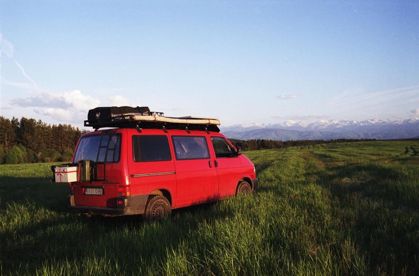 A red van with a roof rack is parked in a grassy field with mountains in the distance.