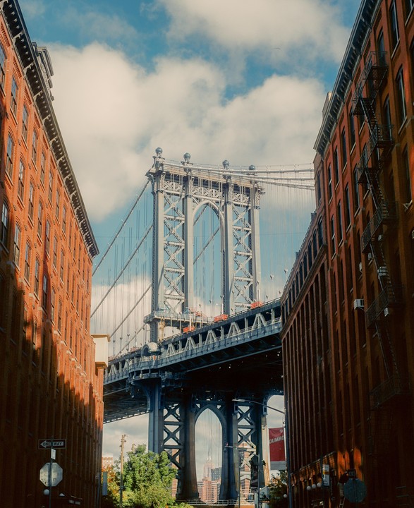 The Manhattan Bridge is framed by two brick buildings under a cloudy sky.
