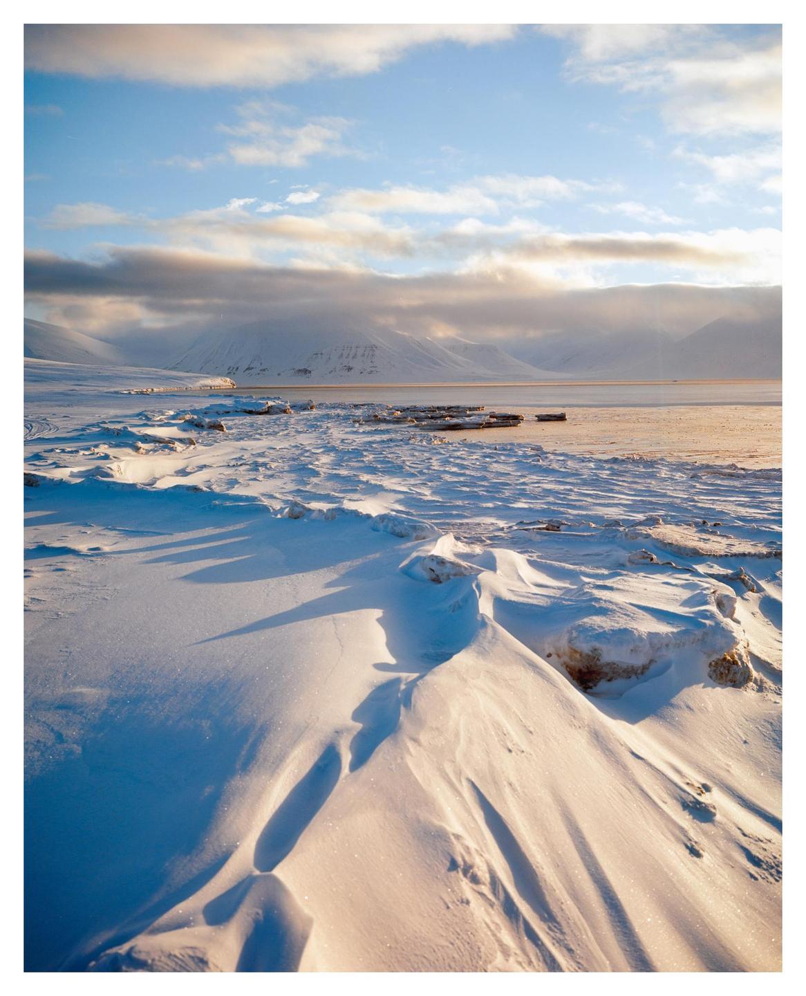 Snow drifts cover the ground with mountains and a body of water in the background under a cloudy sky.