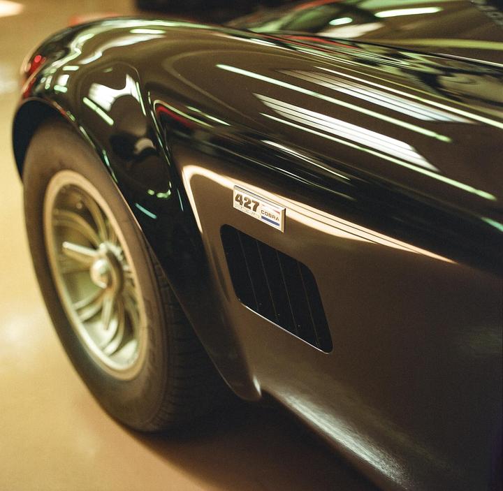 A black Cobra 427 car is parked indoors, with light reflecting off its shiny surface.