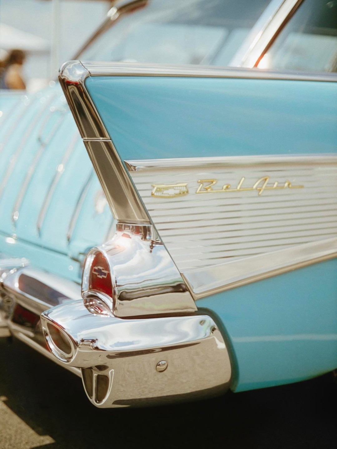 The tail fin and chrome bumper of a light blue vintage car are shown in close-up.