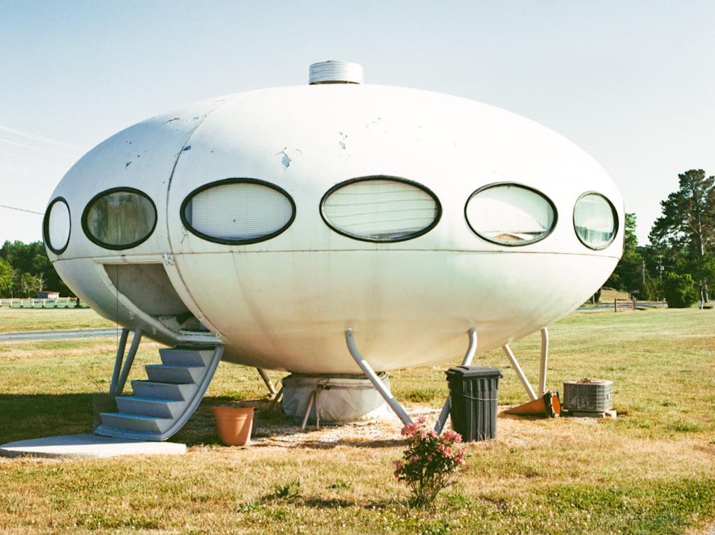 A white, futuristic, flying-saucer-shaped house with oval windows and a staircase leading to its entrance sits in a grassy field under a clear sky.