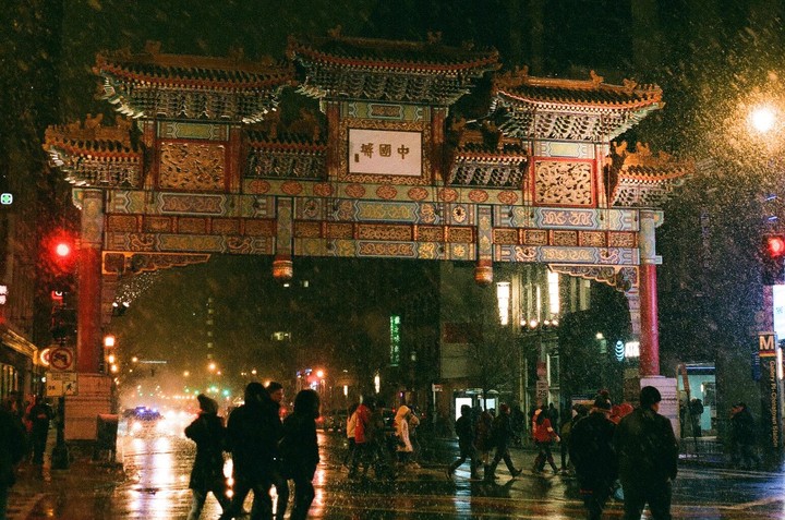 A large, ornate Chinese gate stands over a street at night, with people walking below in the rain.