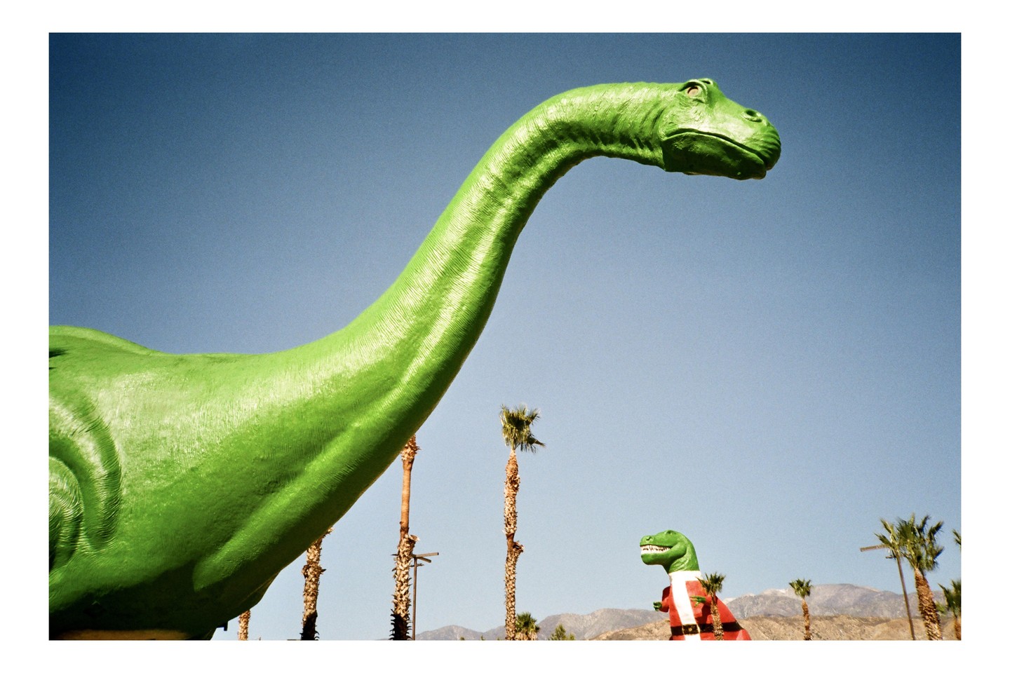 A large green dinosaur statue stands against a clear blue sky, with a smaller dinosaur statue wearing a Santa suit visible in the distance.