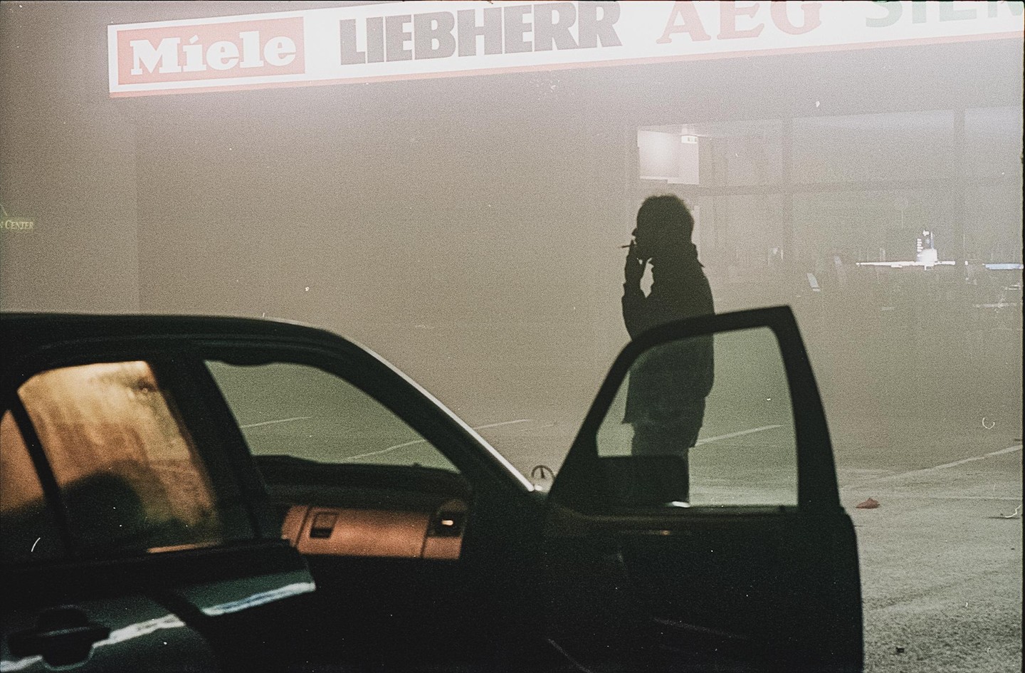 A person smokes a cigarette next to an open car door at night, with a Miele sign visible in the background.