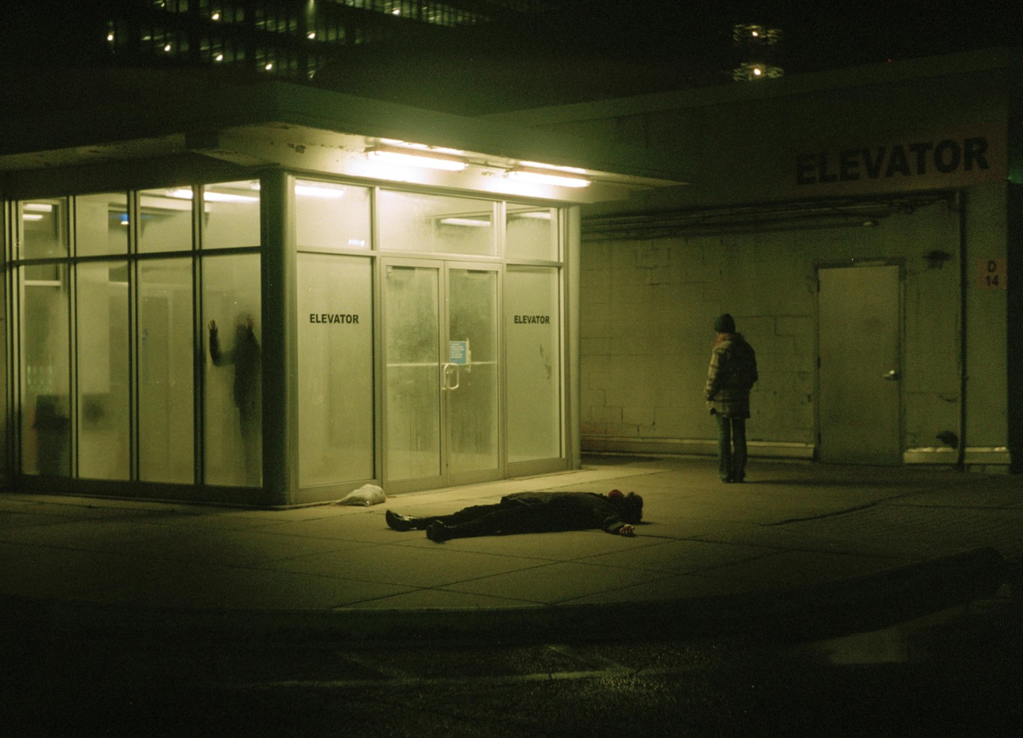 A person lies on the ground in front of an elevator entrance while another person stands nearby at night.