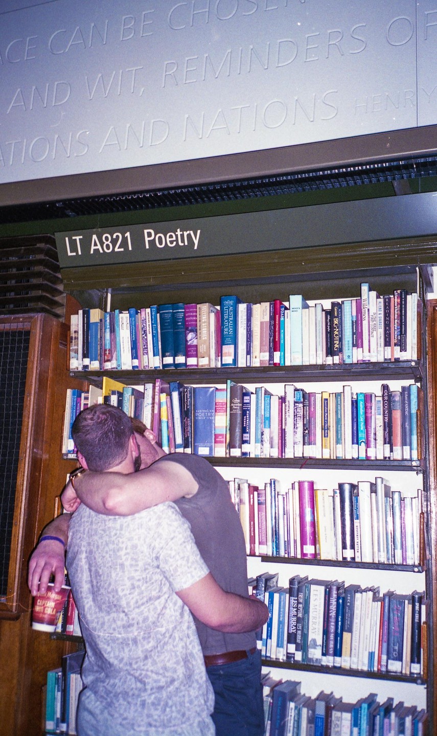 A couple embraces in front of a bookshelf labeled "Poetry" in a library.