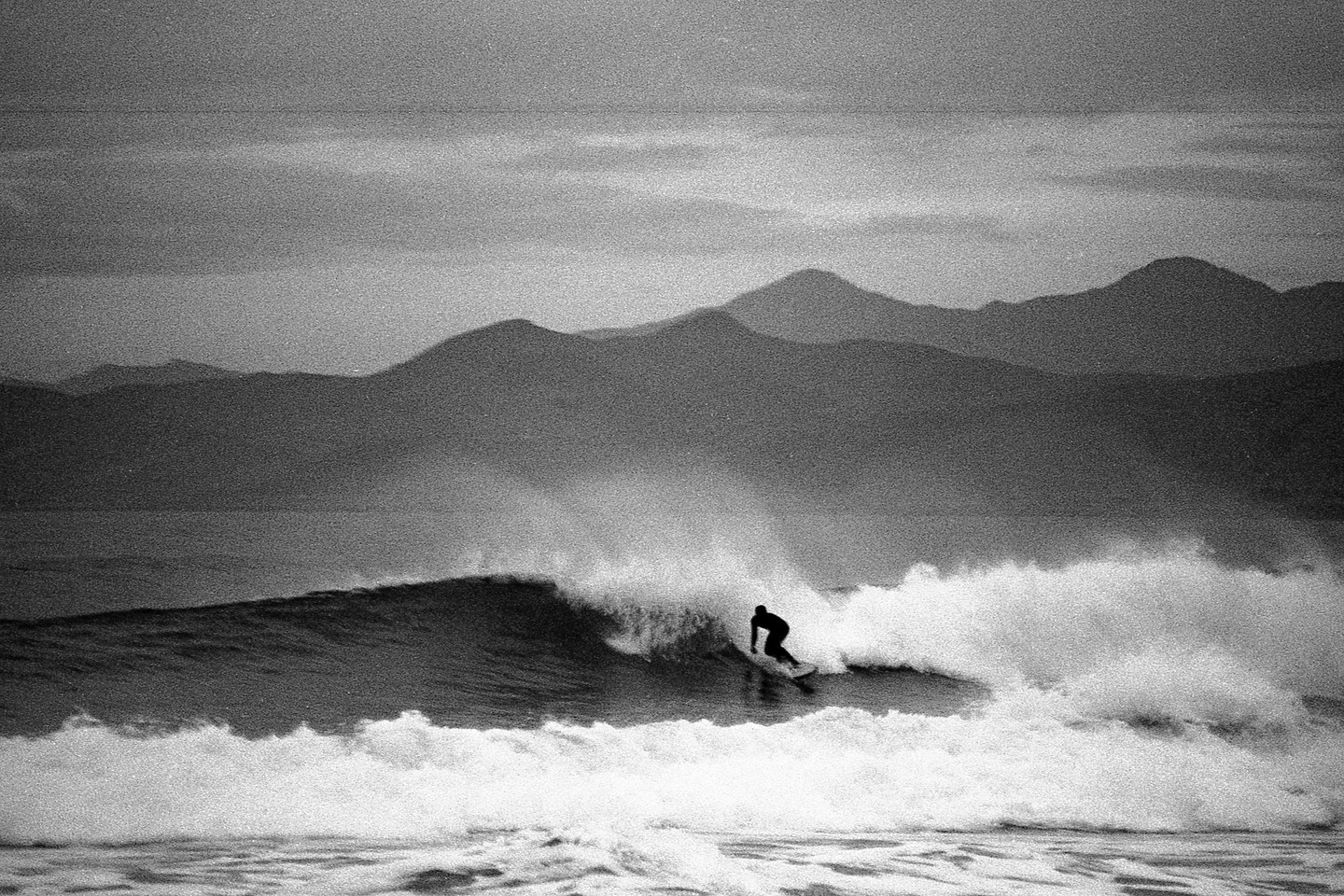 A surfer rides a wave with mountains in the background.
