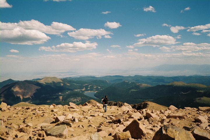 A man holds a child while standing on a rocky mountain peak overlooking a vast landscape of mountains, forests, and a lake under a blue sky with clouds.