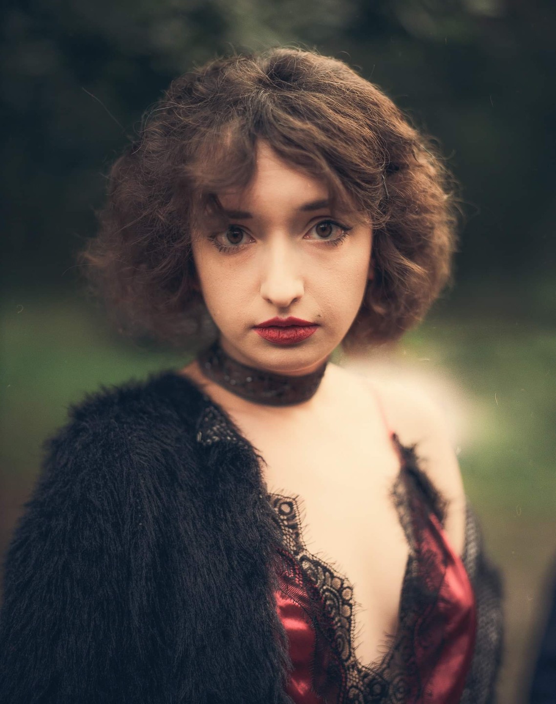 A woman with curly brown hair, red lipstick, and a black choker wears a black furry jacket over a red and black lace top.