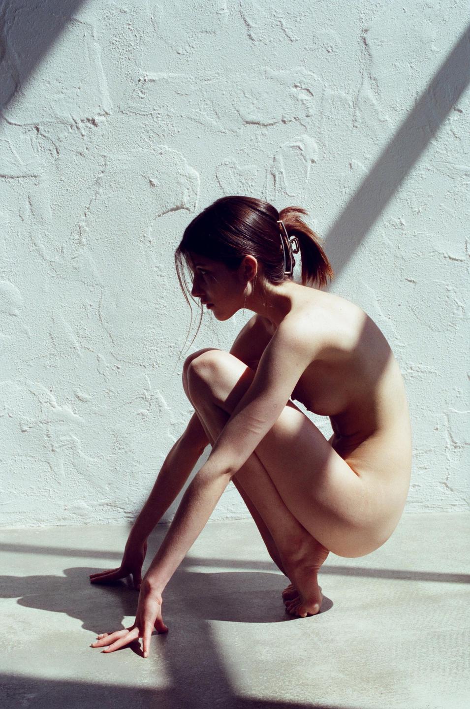 A woman crouches on a floor next to a textured wall with shadows.