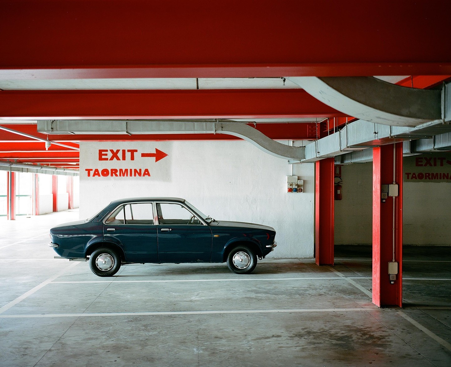 A dark blue car is parked in a garage with red beams and columns, and an exit sign pointing right.